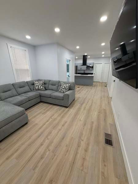 Modern open-plan living room with gray sectional sofa, light wood floor, recessed lights, and kitchen in back