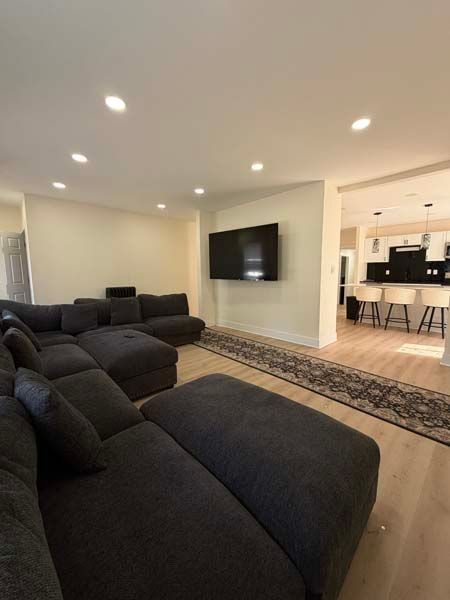 Modern living room with dark sectional sofa, wall-mounted TV, recessed lights, and open kitchen area