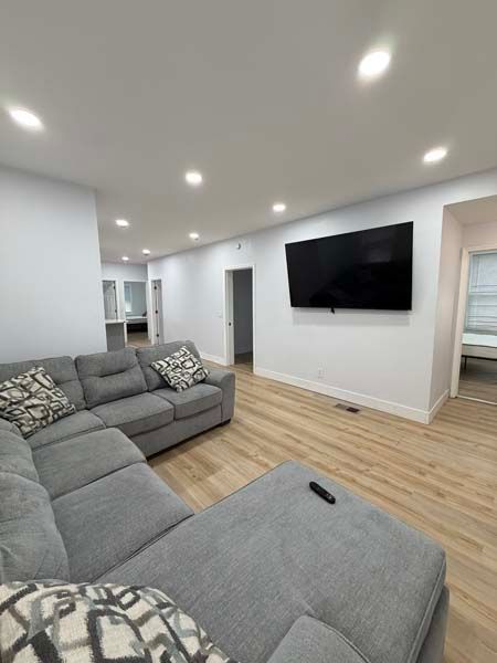 Bright living room with gray sectional sofa, wall-mounted TV, recessed lights, and wood flooring.