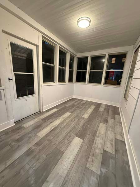 Empty enclosed porch with white walls, windows, and gray wood-look flooring at night.