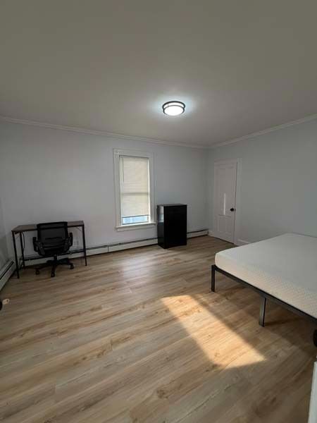 Empty bedroom with light wood floors, white walls, a bed, desk, dresser, and sunlight from a window.