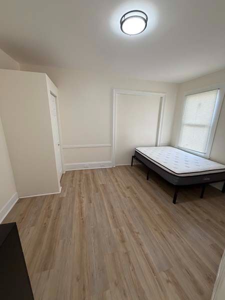 Empty bedroom with light wood floors, white walls, closet, window, and a bed on the right side.