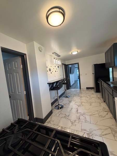 Narrow kitchen hallway with white marble tile, dark cabinets, ceiling lights, and a folded black bed frame in foreground