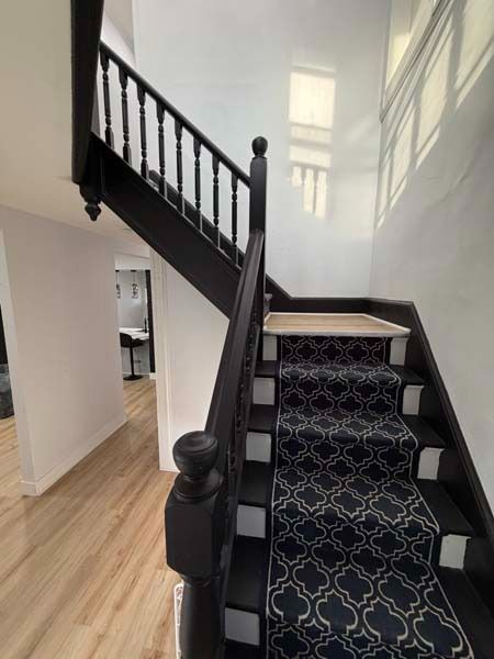 Interior staircase with black railing, patterned carpet runner, and light wood flooring in a bright hallway