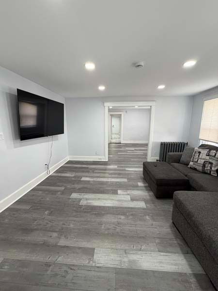 Bright modern living room with wall-mounted TV, gray flooring, recessed lights, and dark sectional sofa