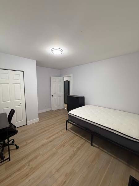 Empty bedroom with a bed, desk chair, and white closet doors on light wood flooring