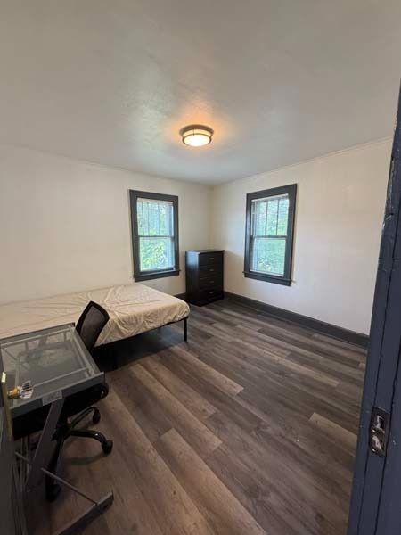Small furnished bedroom with a bed, desk, nightstand, two windows, and wood-look flooring.