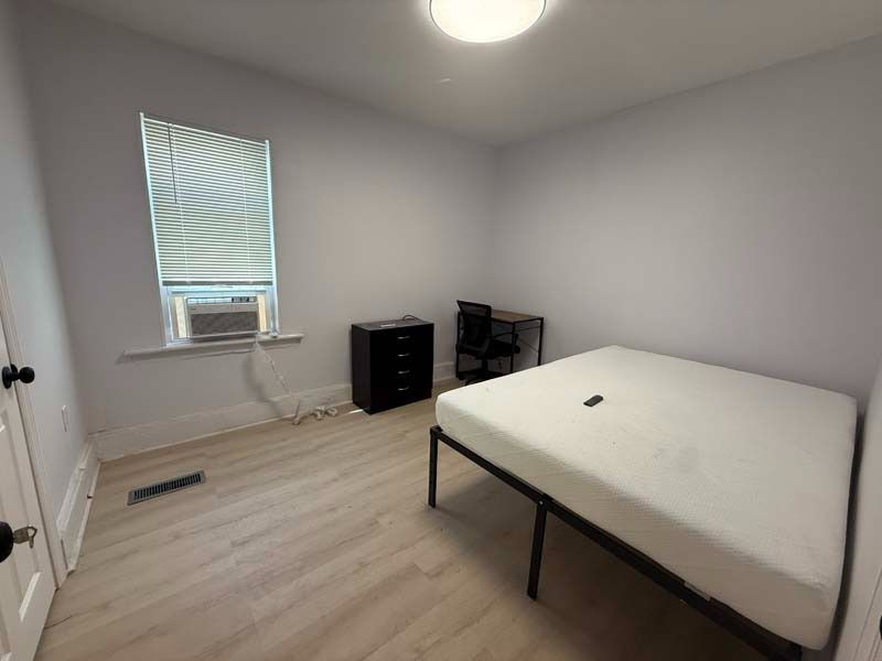 Small bedroom with bed, desk, dresser, window air conditioner, and white walls.