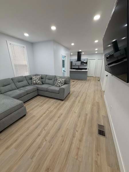 Modern living room with gray sectional, wood floors, white walls, and open kitchen view.