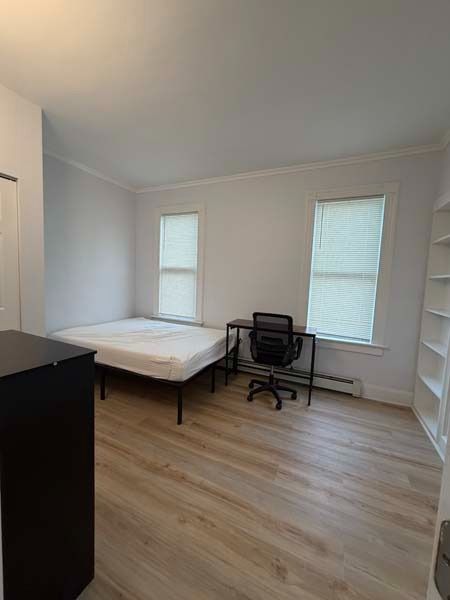 Small bedroom with a bed, desk chair, and two windows on light wood floors.