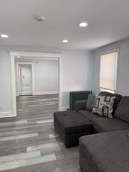 Bright gray living room with a dark sectional sofa, ottoman, radiator, and open doorway to another room