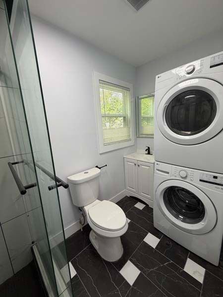 Small bathroom with stacked washer and dryer, toilet, glass shower, and black tile floor.