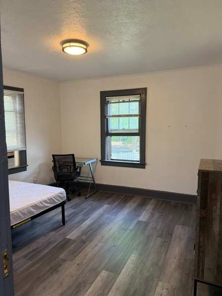 Small bedroom with bed, desk chair, two windows, and dark wood flooring.