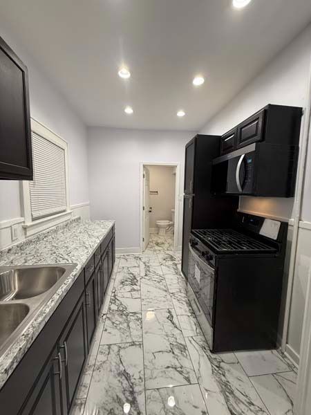 Narrow modern kitchen with white marble floors, dark cabinets, stainless sink, and recessed ceiling lights.