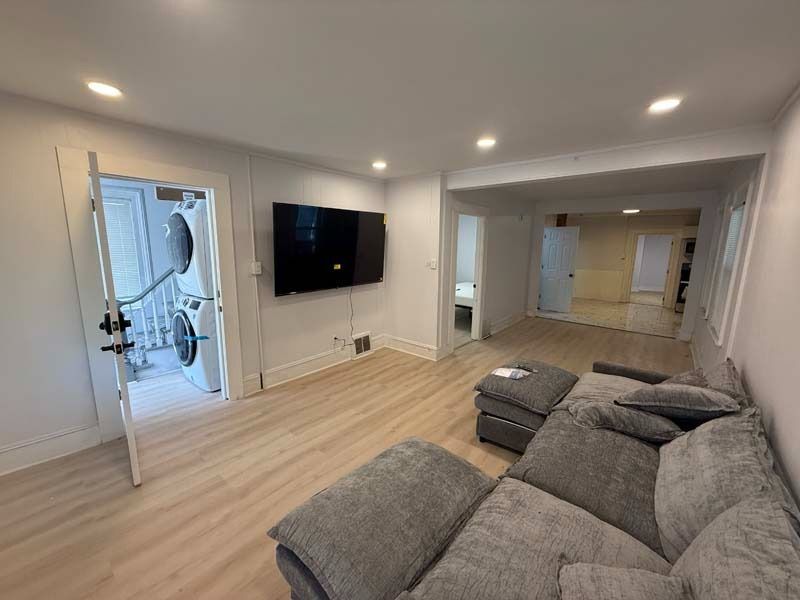 Bright living room with gray sectional, wall-mounted TV, and open front door to hallway
