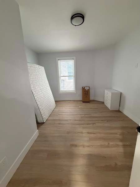 Empty unfurnished room with white walls, wood floor, a mattress leaning against the wall, and a window.