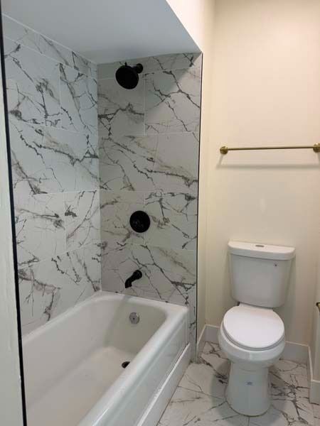 Small bathroom with white tub, marble-look shower wall, and toilet beside a towel bar