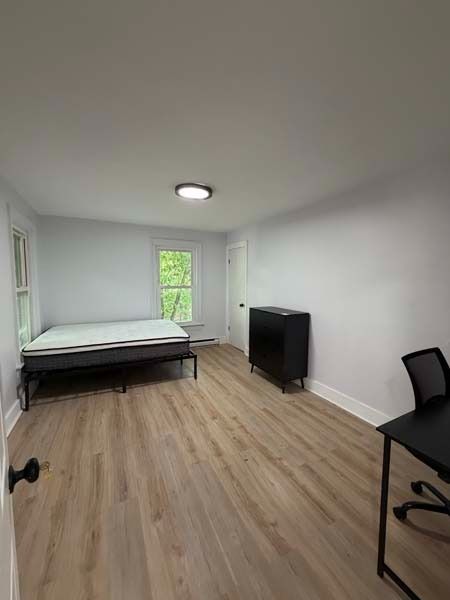 Small furnished bedroom with a bed, desk, chair, dresser, and window on white walls and wood floor