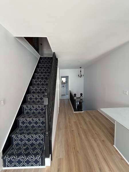Narrow hallway with patterned carpet stairs, light wood floor, white walls, and a bench on the right