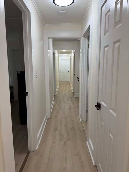 Narrow white hallway with recessed lights and open doors, viewed toward a bright room at the end.
