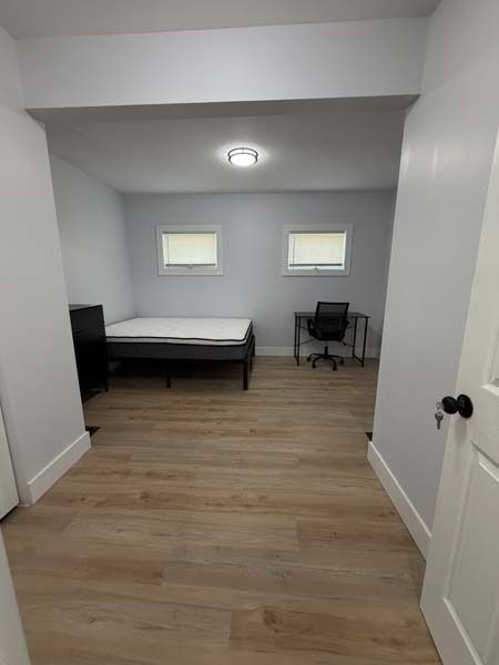 Small furnished bedroom with a bed, desk chair, two windows, and wood flooring.