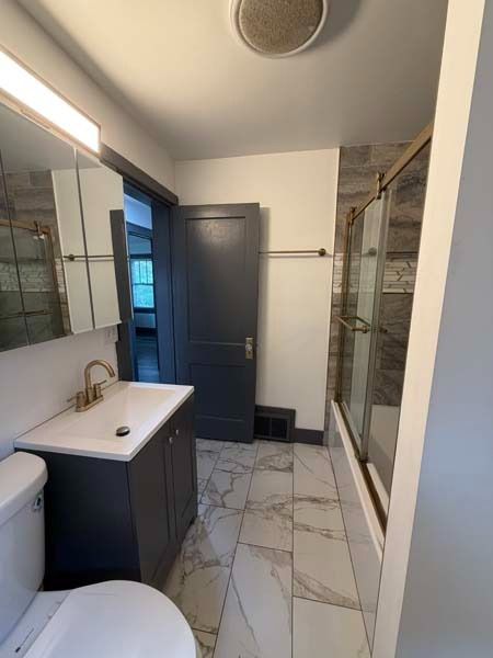 Bright bathroom with white marble floor, dark vanity, mirror, toilet, shower, and gray door