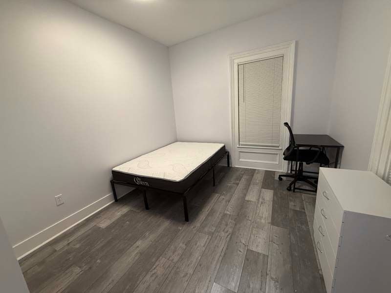 Small furnished room with a bed, desk chair, white walls, and gray wood flooring.