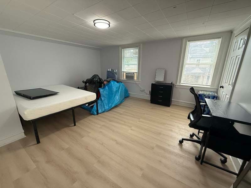Small furnished room with two beds, desk, dresser, windows, and a blue bag against the wall.