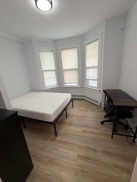 Small bedroom with a bed, desk, chair, and three bay windows with blinds