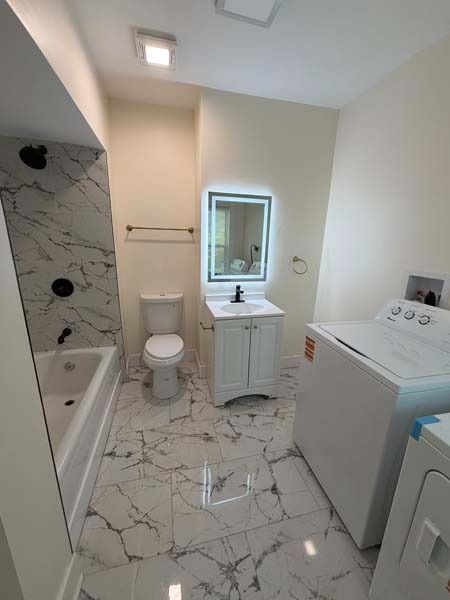 Bright white bathroom with marble tile, tub, toilet, vanity sink, mirror, and a washer/dryer.