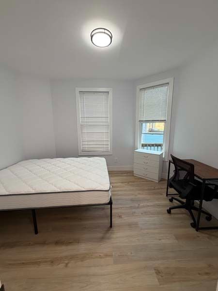 Small bedroom with a bed, desk chair, two windows, white walls, and light wood floors.