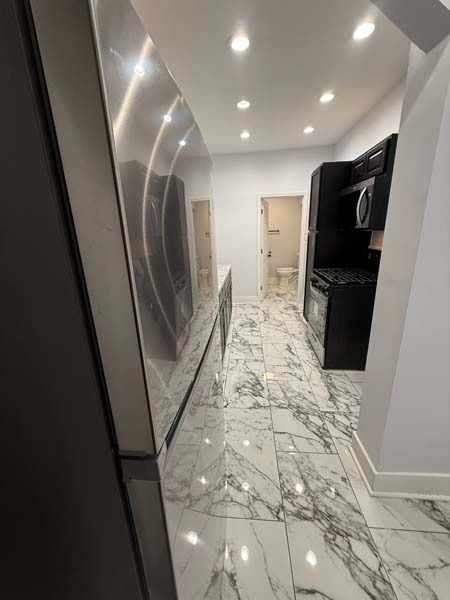 Narrow modern kitchen hallway with glossy marble floor, white walls, recessed lights, and black cabinets.