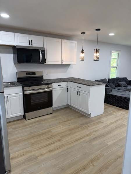 Modern kitchen with white cabinets, stainless steel appliances, gray countertops, and light wood flooring opening to a living room.