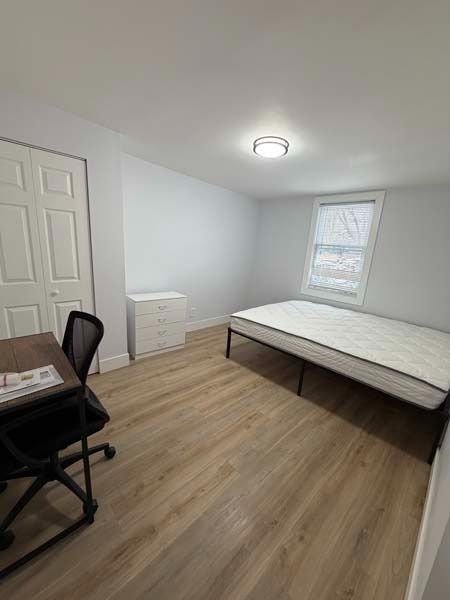 Small furnished bedroom with white walls, wood floors, bed, desk chair, dresser, and window.