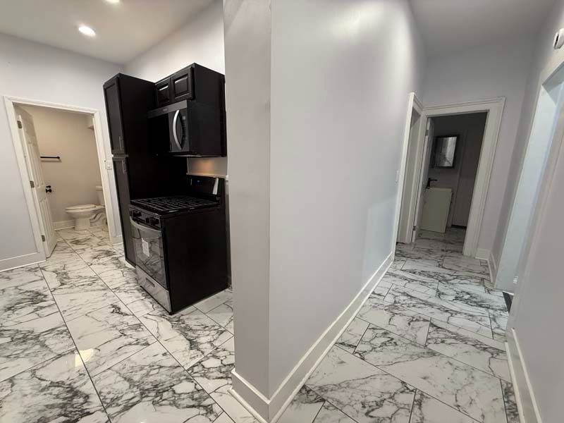 Empty hallway beside a dark kitchen with marble-pattern tile flooring and white walls.