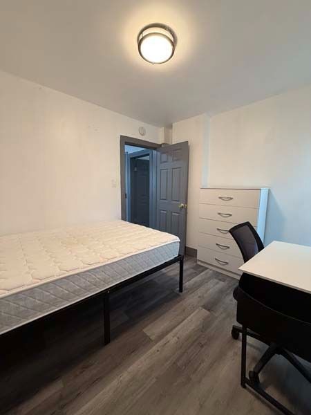 Small bedroom with a bed, dresser, desk, and open doorway, shown in soft lighting.