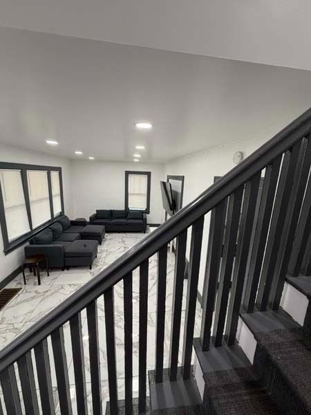 Modern living room with black sectional sofas, large windows, and a staircase railing in the foreground