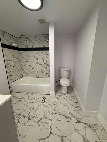 Small bathroom with marble-pattern tile, bathtub on left, toilet in separate alcove, and plain white walls