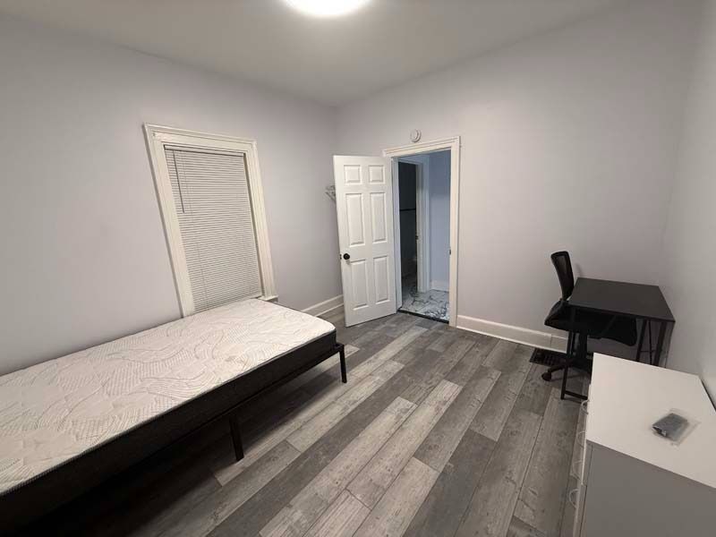 Spare bedroom with twin bed, desk, chair, and open door on gray wood flooring.