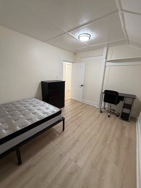 Small furnished bedroom with bed, dresser, desk, and chair under a ceiling light.