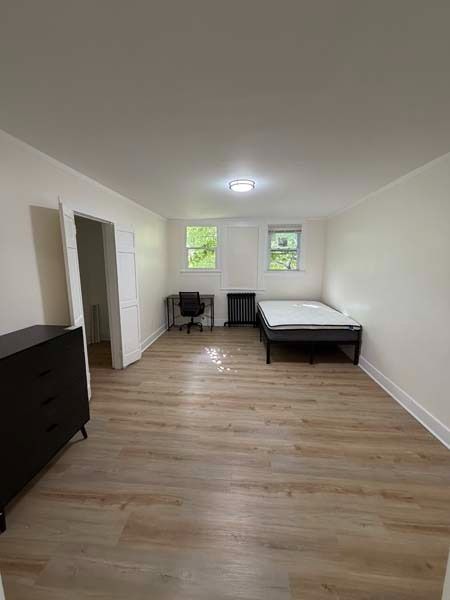 Spare bedroom with bed, dresser, desk, and two windows on light wood floors.