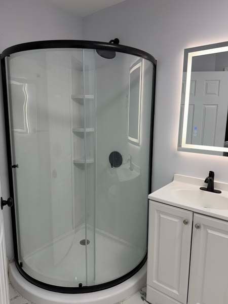 Curved glass shower stall with black trim beside a white vanity and lit mirror in a bathroom