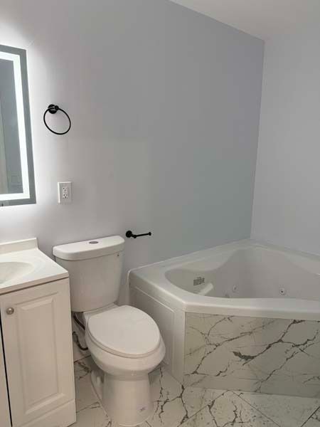 Small bathroom with white vanity, toilet, and marble corner soaking tub against light gray walls