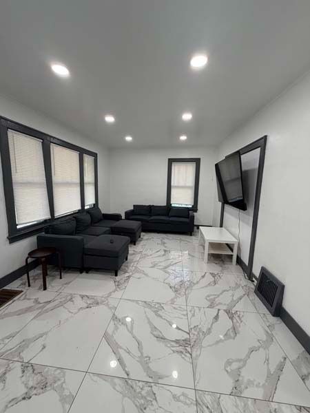 Modern living room with black sofas, wall-mounted TV, marble tile floor, and bright recessed ceiling lights