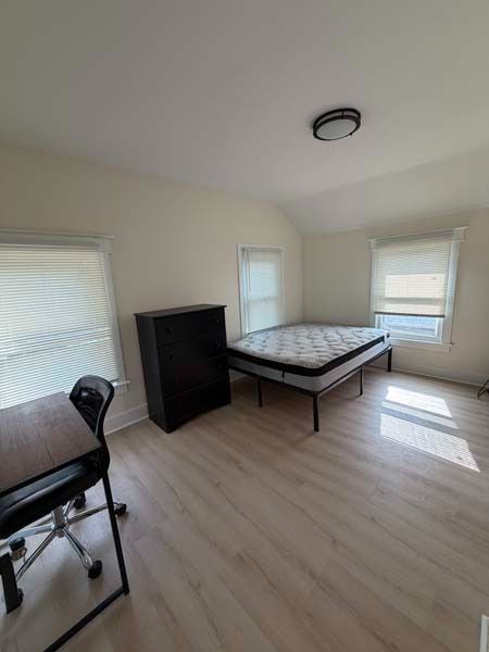 Empty bedroom with bed frame, dresser, and exercise bike, bright white walls and blinds
