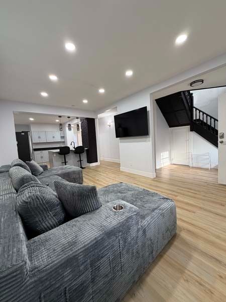 Bright open living room with gray sectional sofa, wall-mounted TV, wood floors, recessed lighting, and staircase.