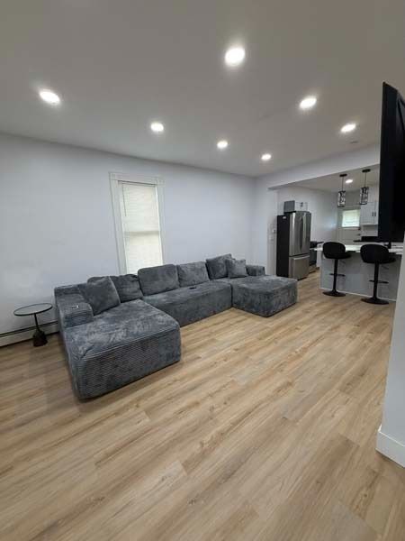 Bright modern living room with gray sectional sofa, wood floors, recessed lights, and a kitchen bar in the background