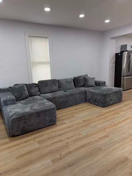 Modern living room with gray sectional sofa, light wood floor, recessed lights, and stainless-steel refrigerator