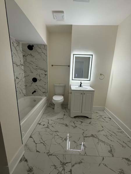 Small bathroom with marble-pattern tile, bathtub, toilet, vanity sink, and mirror.