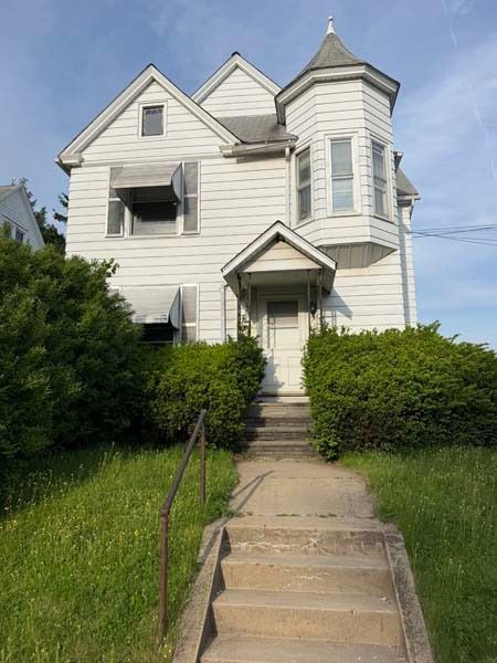 White house with a steep roof and front steps, framed by overgrown hedges and a concrete walkway